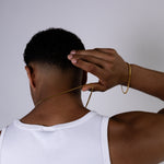 A man viewed from behind wearing a white ribbed tank top, demonstrating a 4mm gold chain necklace draped over his neck and a matching gold 4mm chain bracelet on his wrist against a plain light grey background.
