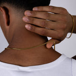 A man viewed from behind wearing a white ribbed tank top, demonstrating a 4mm gold chain necklace draped over his neck and a matching gold 4mm chain bracelet on his wrist against a plain light grey background.